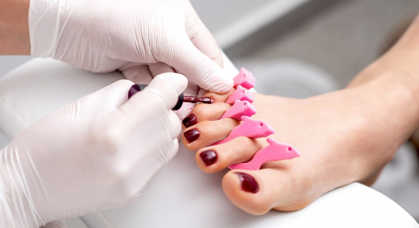 manicure-master-is-painting-female-toenails-with-maroon-nail-polish-by-brush-wearing-white-gloves manicure-master-is-painting-female-toenails-with-maroon-nail-polish-by-brush-wearing-white-gloves